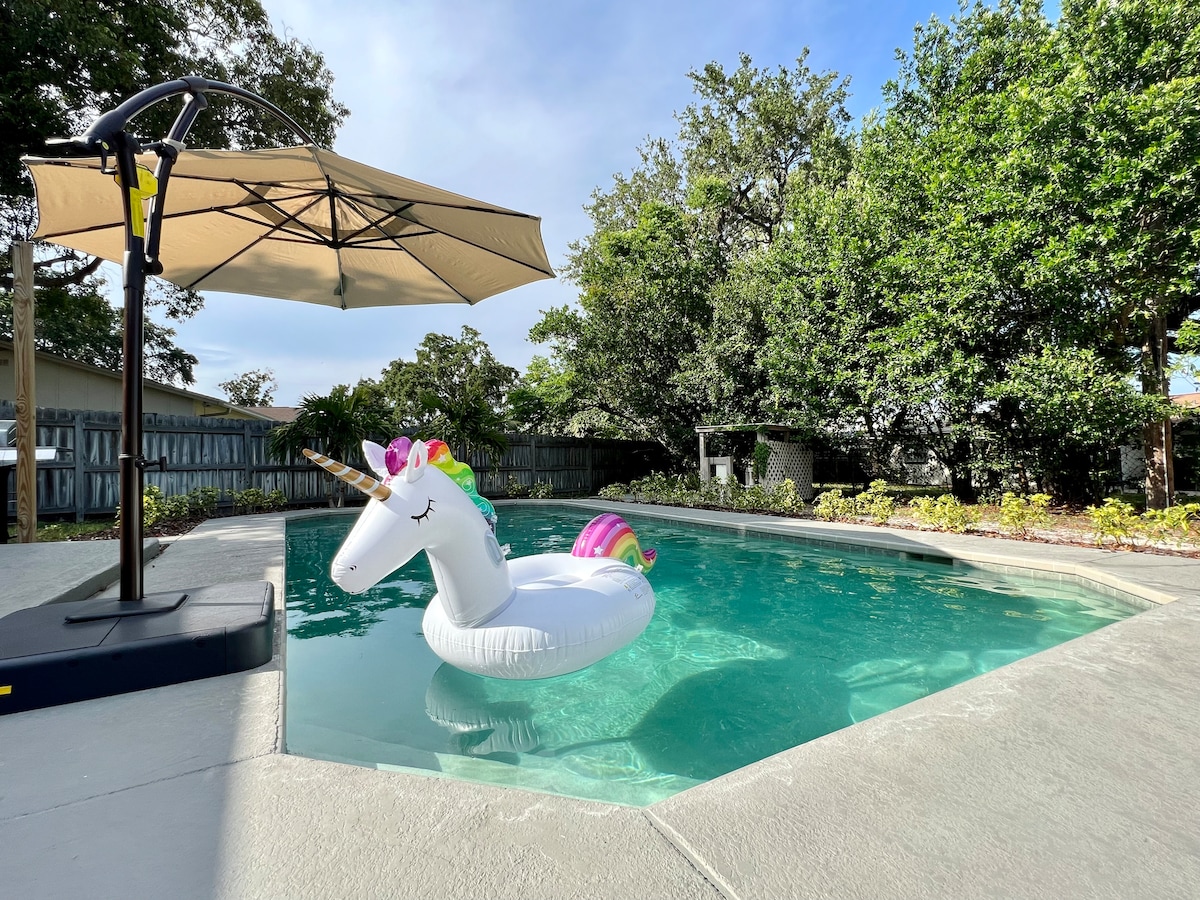 A private in-ground pool is surrounded by lush landscaping, featuring a unique unicorn float. A shaded umbrella stands nearby, offering a cozy spot to relax while enjoying the tranquil outdoor setting.