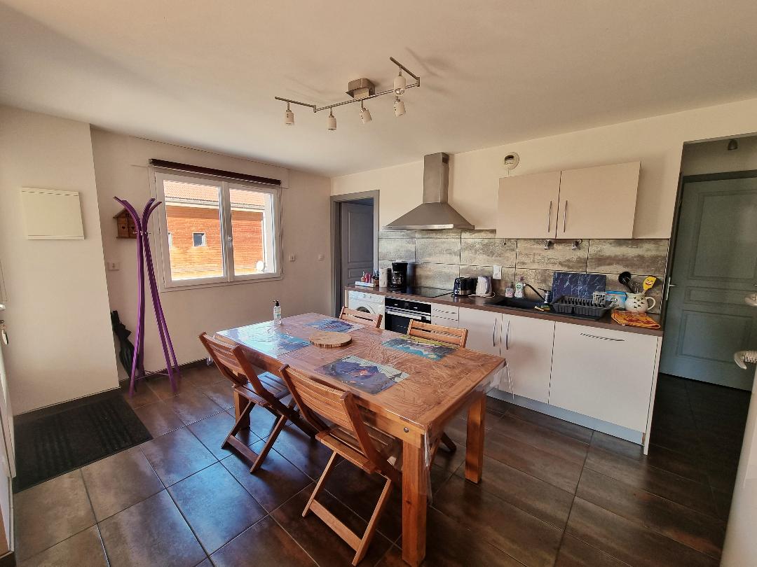 A modern kitchen features a wooden dining table surrounded by six chairs. A sleek range hood is positioned above the stove, and appliances are arranged along the countertops. Natural light enters through a window, illuminating the workspace.