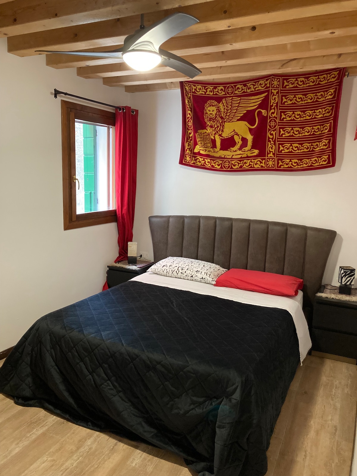A comfortable bedroom is highlighted by a spacious bed with a dark quilt. Accent pillows in red and white complement the bedding. A wooden ceiling with exposed beams adds character. A colorful flag hangs above the bed, and natural light enters through the window, framed by red curtains.