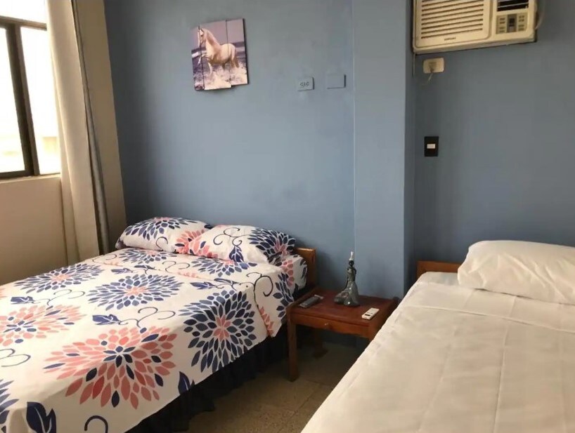 A light blue room contains two beds, one with a floral-patterned bedding set. A nightstand holds a small decorative piece. Natural light enters through a large window, and a wall-mounted air conditioning unit is visible.