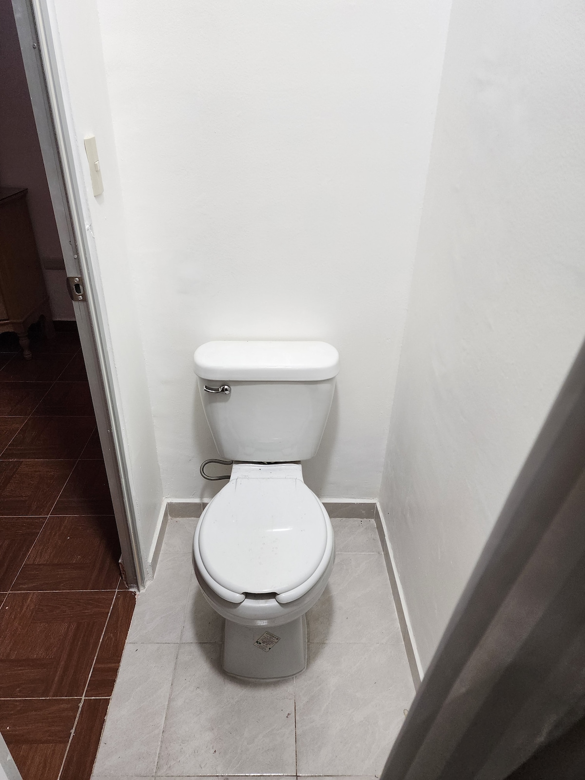 A clean and functional bathroom space is depicted, featuring a white toilet against a plain white wall. The flooring is tiled in light grey and brown, with a small wooden cabinet visible through the open door.