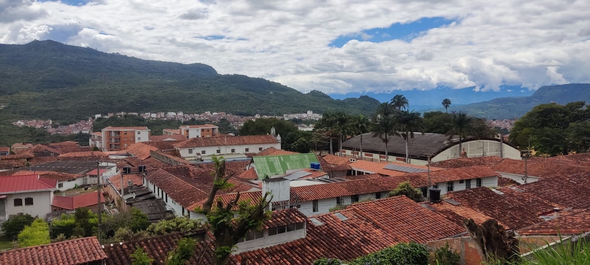 A panoramic view of a vibrant neighborhood is displayed, featuring terracotta roofs and lush greenery. The backdrop is dominated by rolling hills and a partly cloudy sky, while the distant mountains add depth to the scenic landscape.