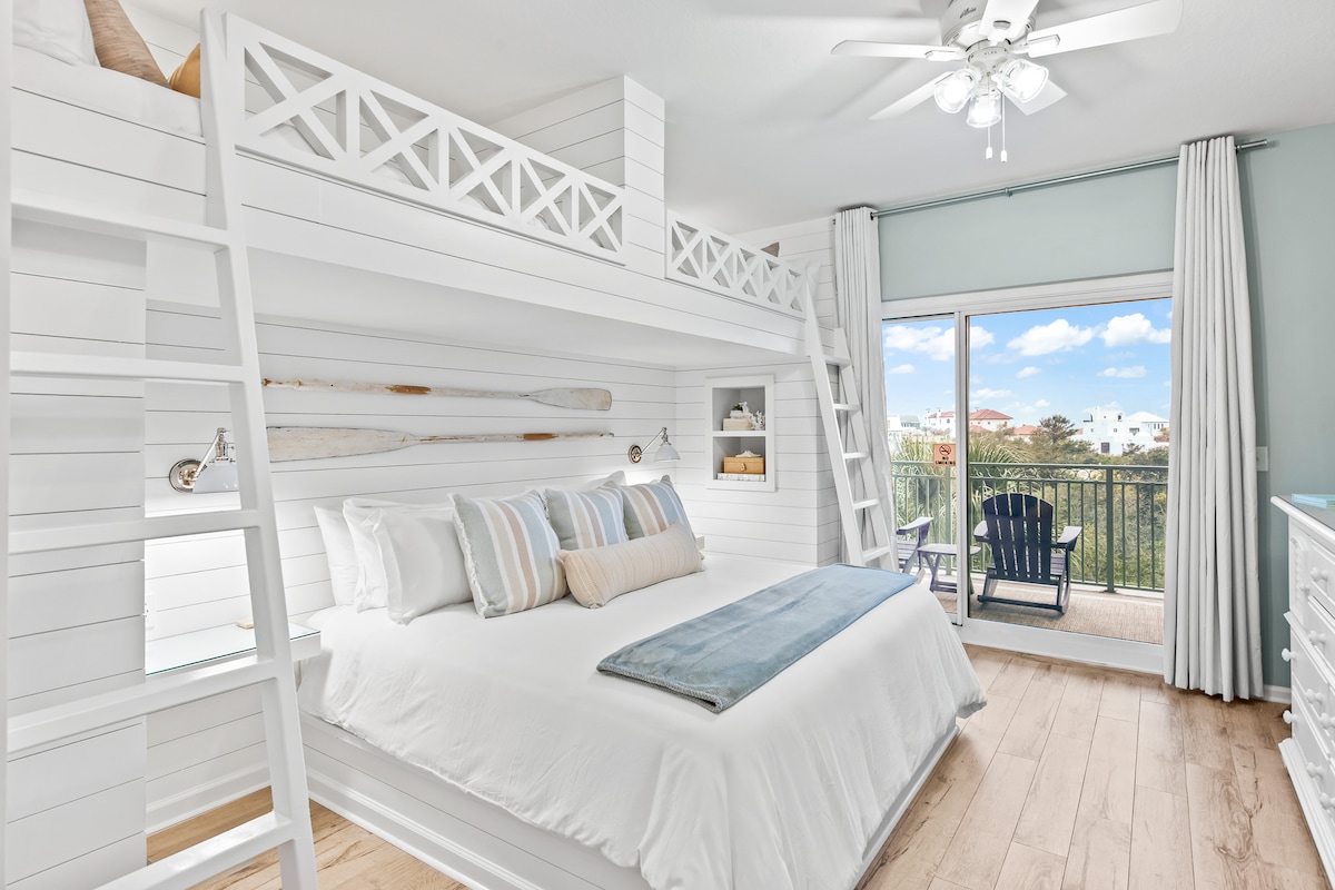 The sleeping area features a spacious bed with light-colored bedding, complemented by a soft throw. Above, a double twin bunk setup is visible, enhancing practicality for families. Large windows allow natural light, and a balcony with chairs overlooks the surrounding area.