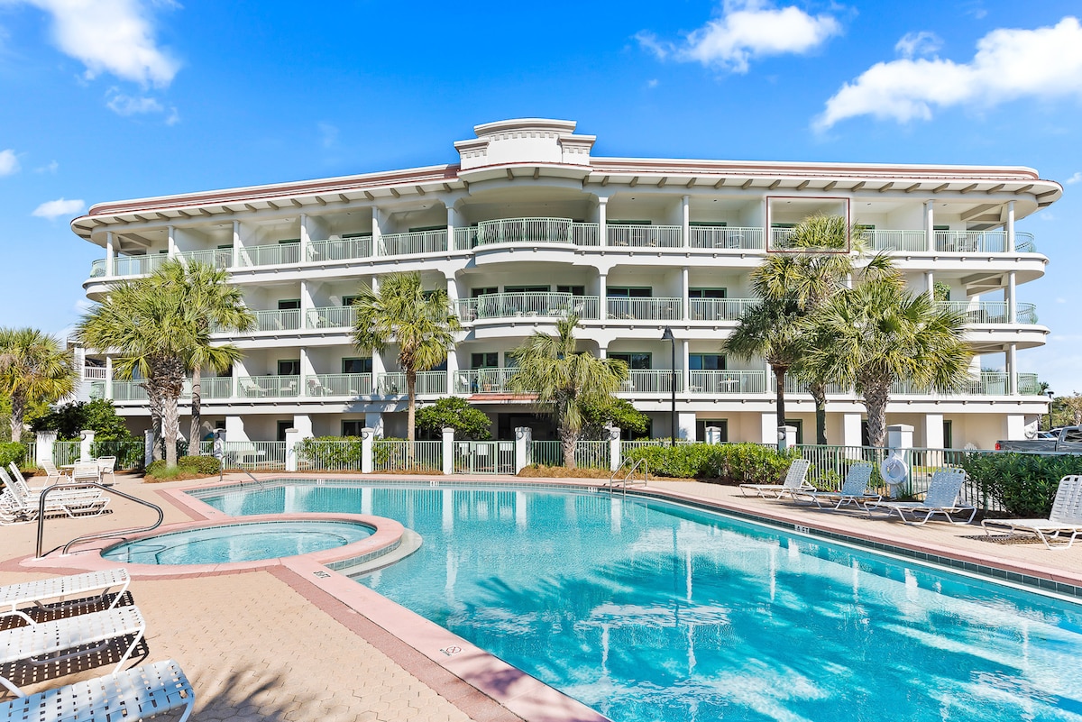 A modern four-story condo complex is visible, featuring multiple balconies overlooking a spacious pool. Palm trees surround the area, and lounge chairs are neatly arranged around the pool. Clear skies enhance the inviting atmosphere of the outdoor space.