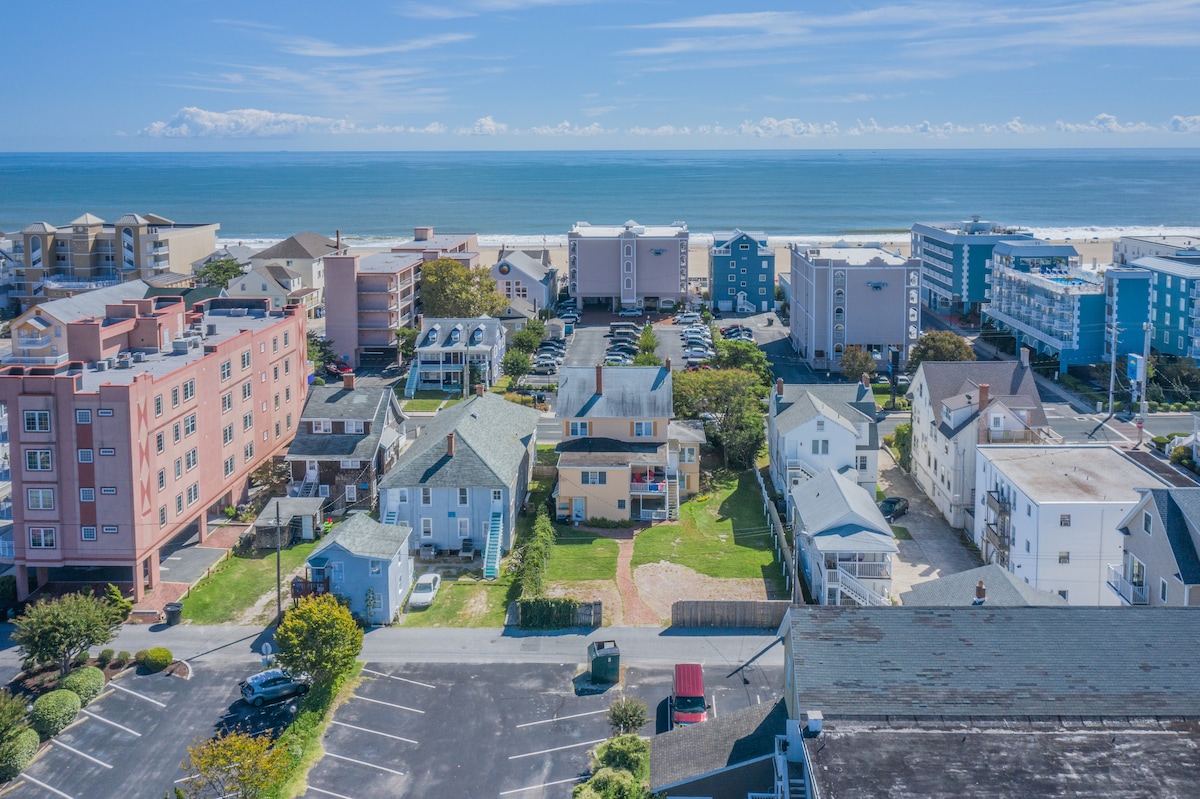An aerial view captures a mix of residential and commercial buildings near the beach. The coastline extends in the distance, with clear skies overhead. Several parking areas and green spaces are visible, contributing to the community atmosphere.