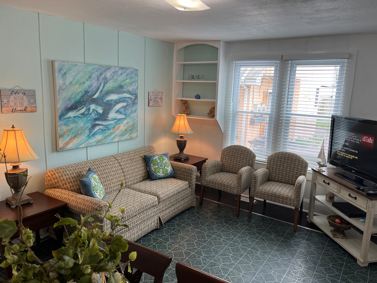 A cozy living area is arranged with a comfortable sofa and two accent chairs. A large piece of artwork adorns the wall, and two lamps provide soft lighting. The space is brightened by natural light streaming through the windows, enhancing the inviting atmosphere.