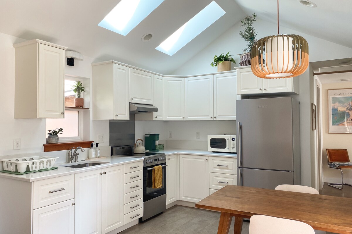 A bright kitchen is showcased with white cabinetry and skylights above. Modern appliances, including a stainless-steel refrigerator and oven, are visible. A dining table for two adds a cozy touch, while plants are observed on the shelves, enhancing the fresh atmosphere.