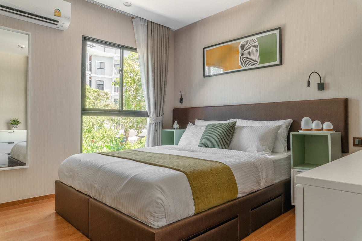 A modern bedroom features a spacious bed with a soft, neutral duvet and an accent green throw. Natural light streams through a window, illuminating the room, while accent artwork and bedside tables add detail. A large mirror reflects the inviting atmosphere.