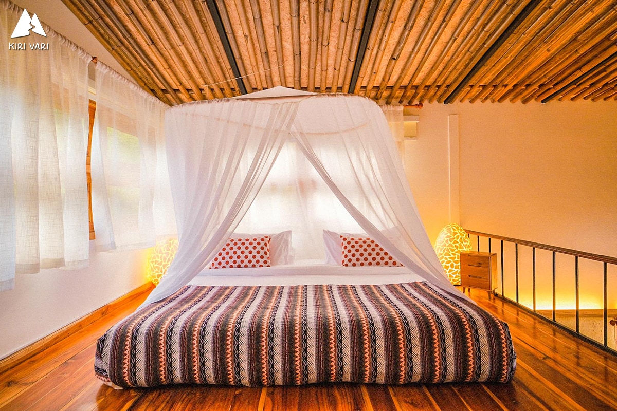 A spacious bed is centered in the room, draped with a white canopy. Textured wooden flooring complements the natural bamboo ceiling, while soft lighting creates a calming atmosphere. Two decorative pillows with red patterns are arranged at the head of the bed, enhancing the serene space.