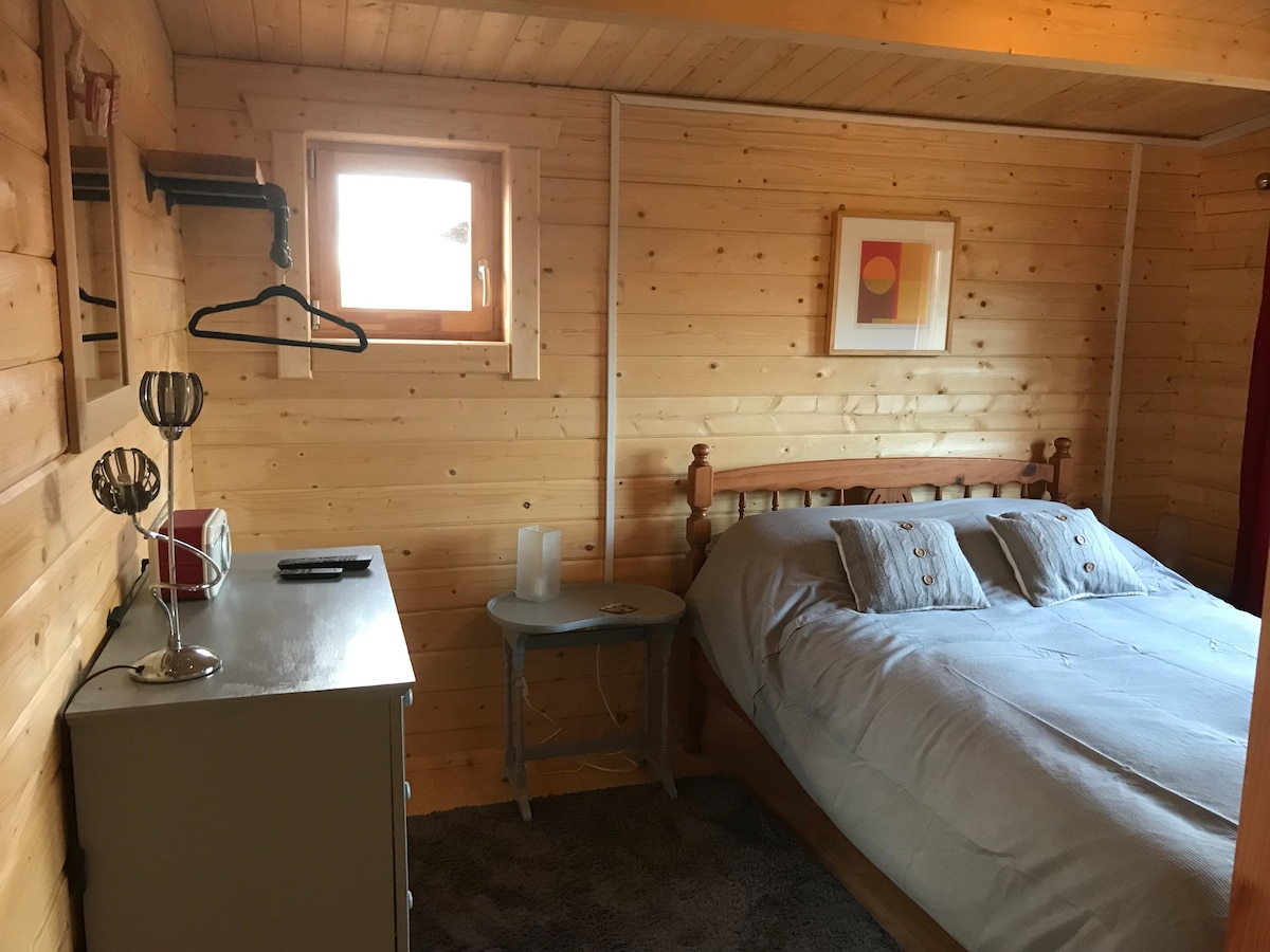 A cozy interior features natural wood paneling and a spacious bed with soft linens. A bedside table and a drawer unit provide storage, while a small table and a floor lamp add functionality. A window brings in natural light, creating a warm ambiance.