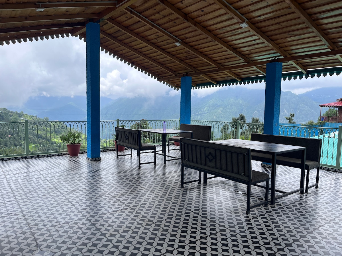 A spacious terrace features a patterned tiles flooring with several black wooden tables and benches positioned to offer views of the surrounding mountains. Lush greenery and distant hills are visible under a cloudy sky, creating a serene atmosphere for outdoor relaxation.