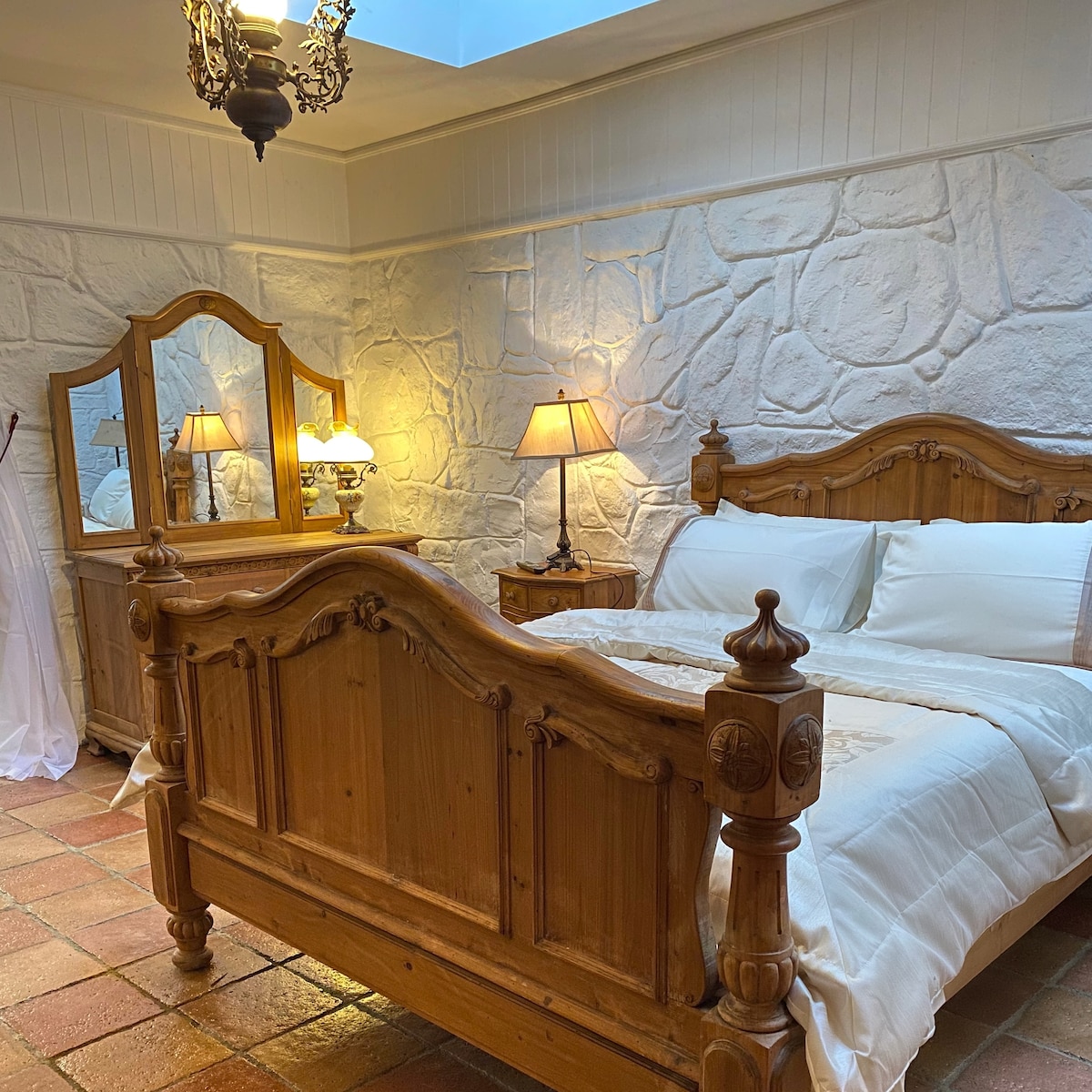 A spacious bedroom is highlighted by a wooden bed frame with an intricate design. Soft light from a bedside lamp illuminates the light-colored walls, complemented by a stone texture. An elegant mirror is positioned above a wooden dresser, adding functionality to the serene atmosphere.
