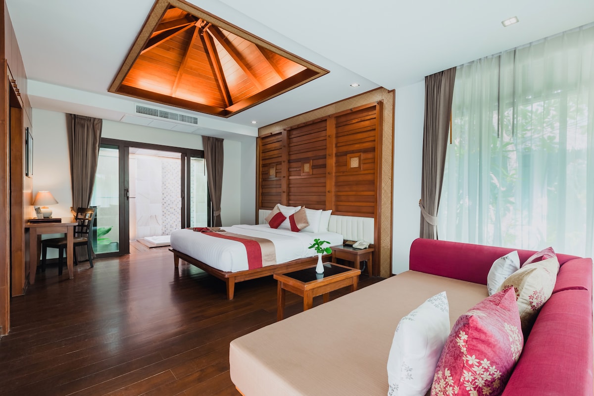 A spacious guest room features a large bed with crisp white linens and a modern design. Natural light enters through wall-to-wall windows, illuminating wooden accents and soft furnishings. A cozy seating area is provided, styled with colorful cushions. A balcony is visible in the background.