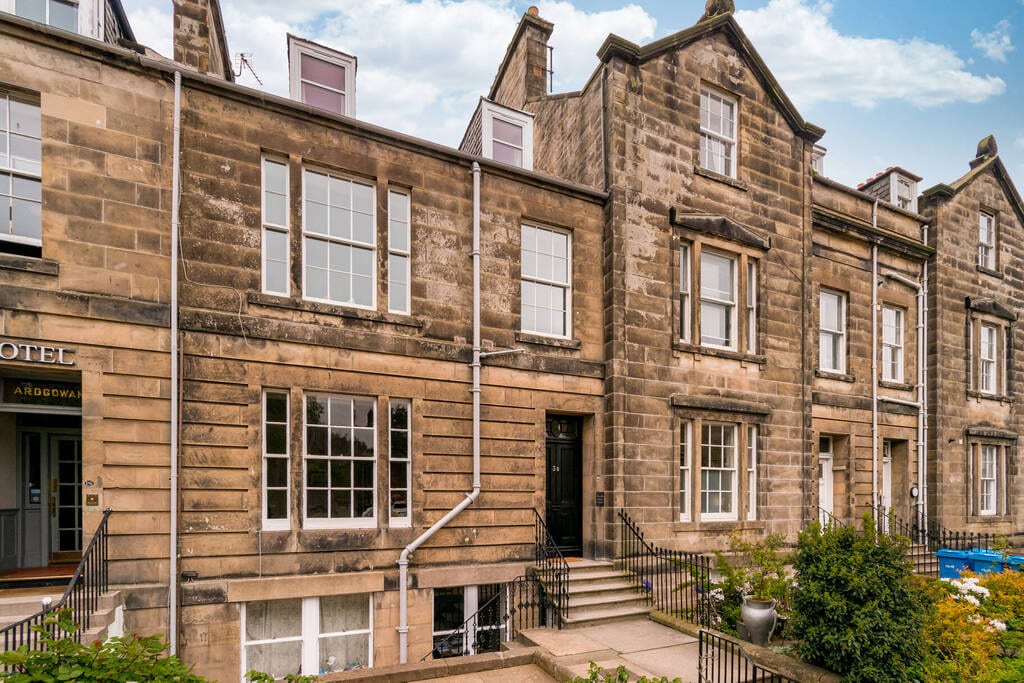 The exterior of a historic stone building is showcased, featuring a symmetrical façade with large windows and distinctive architectural details. A set of stone steps leads to the main entrance, framed by manicured greenery and a well-kept pathway.