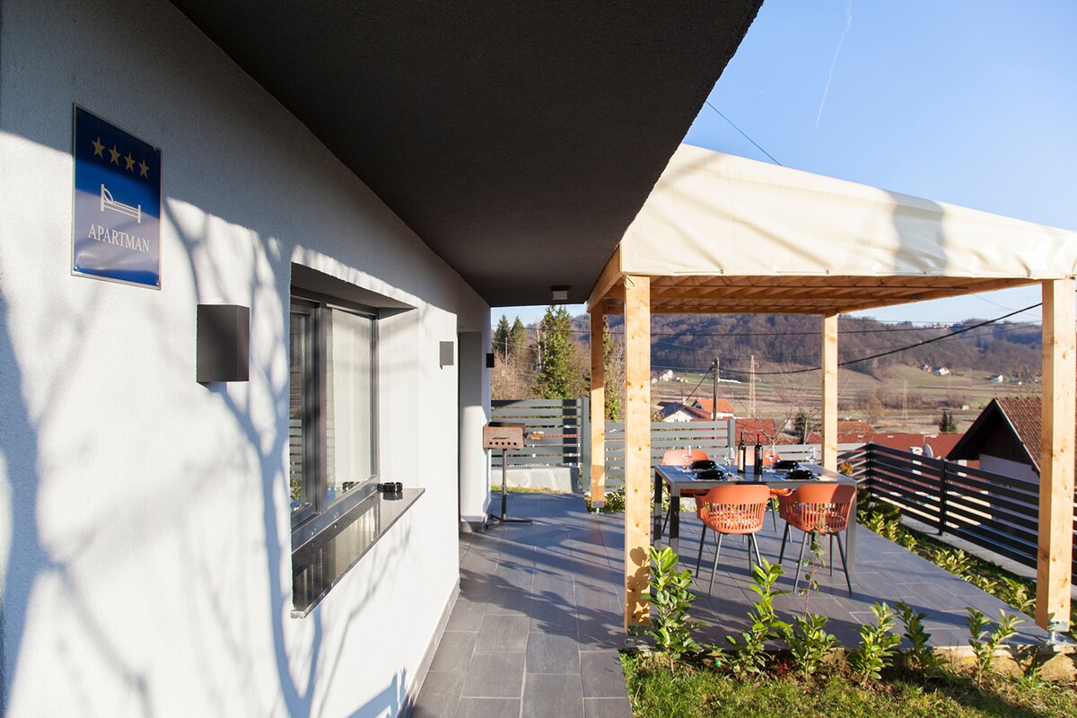 An outdoor terrace features a wooden pergola, providing shade for a dining area with a table and chairs. A barbecue grill is situated nearby. The open view showcases the surrounding landscape, with clear skies and distant hills visible in the background.