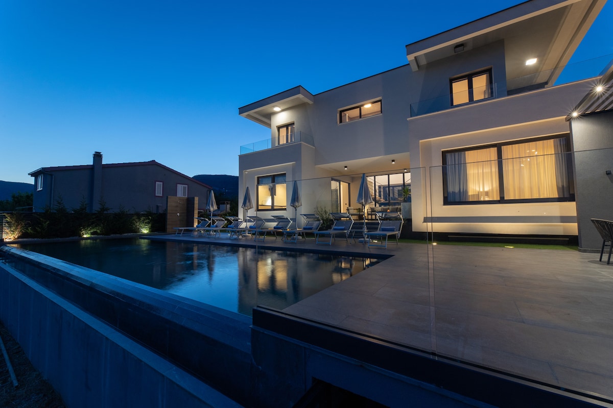 A modern villa is shown at dusk, featuring a sleek pool reflecting the evening sky. Lounge chairs are arranged around the pool area, and large glass doors provide a glimpse into the interior spaces, highlighting a connection between inside and outside.