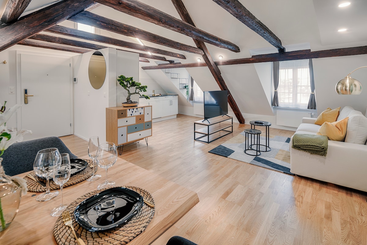 An open living space features high wooden beams that highlight the room's architecture. A modern sofa is paired with a minimalist coffee table. A dining area is set with glassware on elegant plates. Natural light floods in through the large windows, creating an inviting atmosphere.