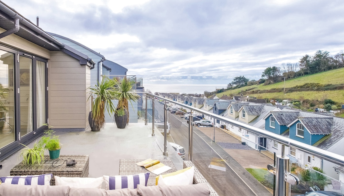 An outdoor balcony features modern furniture with cushions, offering a view of the ocean and surrounding hills. Potted plants add greenery, while a clear railing provides an unobstructed sightline of the neighborhood below and the sea horizon.