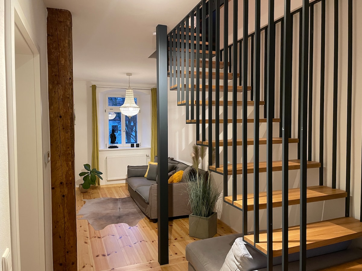 A modern staircase with wooden steps and a decorative iron railing leads to an upper level. Below, a spacious living area is visible, featuring grey sofas, a soft rug, and a plant. Large windows allow natural light to illuminate the neutral walls, complemented by warm curtains.
