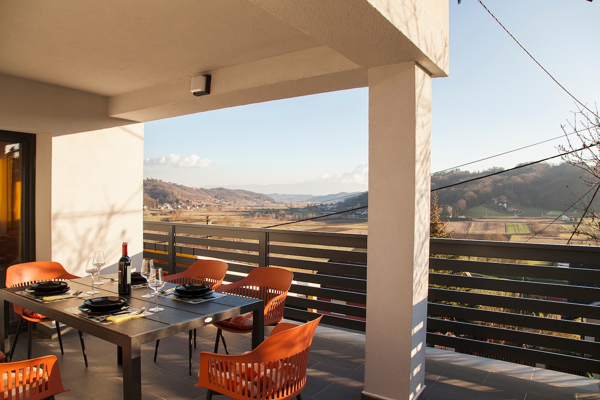 An outdoor terrace features a dining table set for six, surrounded by vibrant orange chairs. Scenic views of valleys and hills are visible in the background, complemented by a clear sky. Soft lighting enhances the space for evening dining.