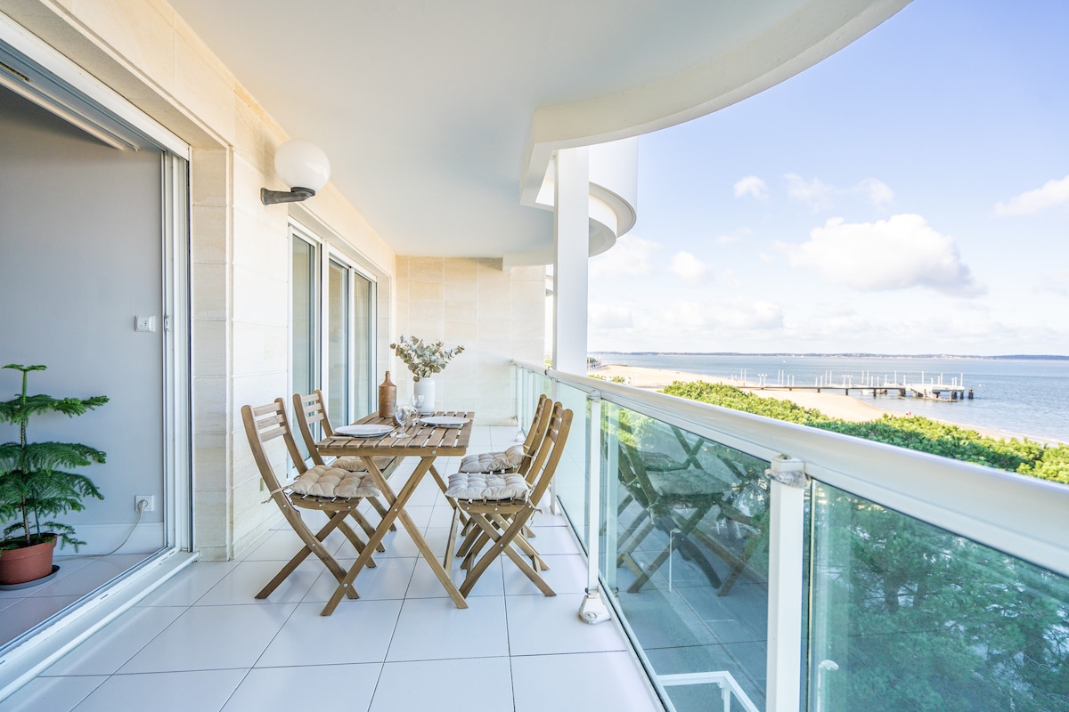 An outdoor terrace includes a wooden dining table with matching folding chairs, set against a backdrop of expansive water views. Sunlight illuminates the space, while a decorative plant adds a touch of greenery to the area. The scene is framed by glass railings.