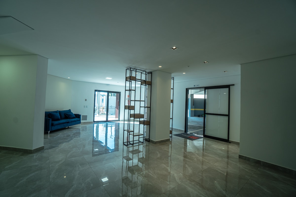 A spacious lobby area features modern design elements with polished marble flooring. A seating area with a blue sofa is positioned against a wall. Large glass doors lead to an outdoor space, allowing natural light to fill the interior.