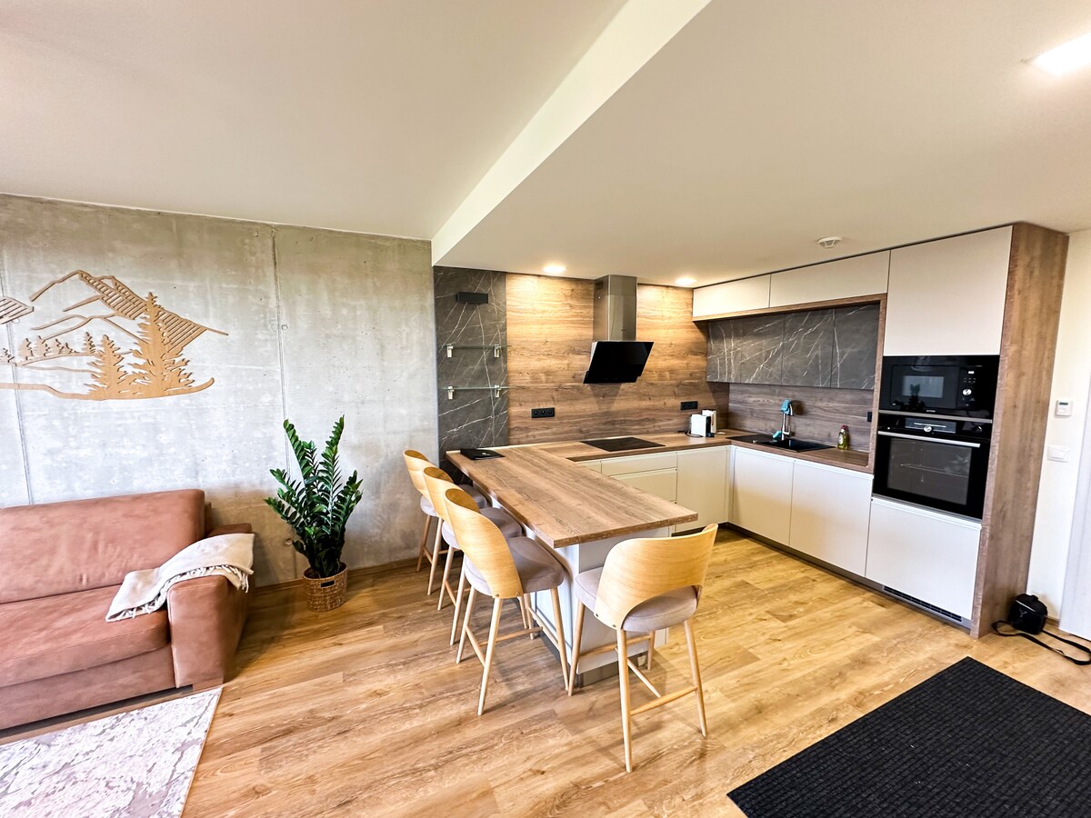 A modern kitchen is presented with sleek cabinetry and a central island featuring six wooden chairs. Appliances include an oven and a stovetop, with textured wall elements adding depth. A comfortable brown sofa sits nearby, complementing the spacious and functional layout.