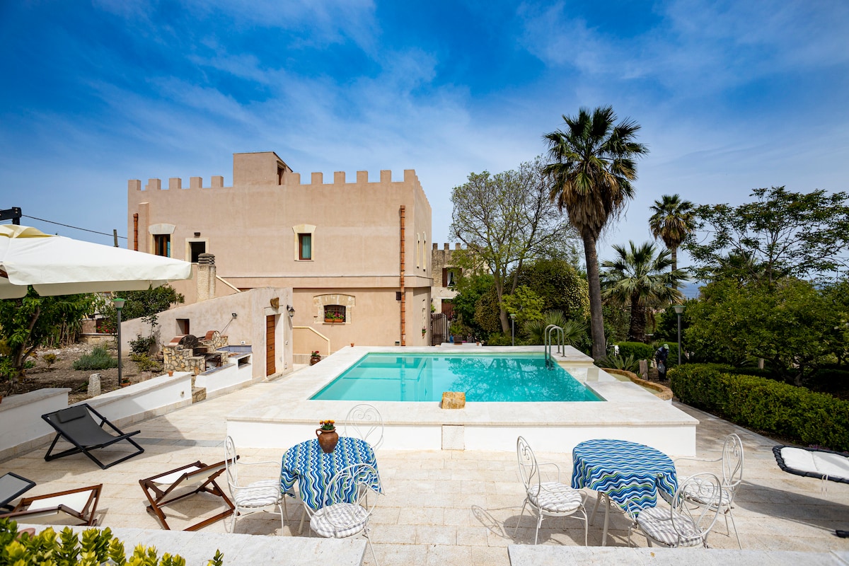 A private pool reflects the clear sky, surrounded by a sun terrace with lounge chairs and outdoor seating. The historic structure features a rectangular layout, accented by lush greenery and palm trees, creating a serene outdoor space for relaxation.