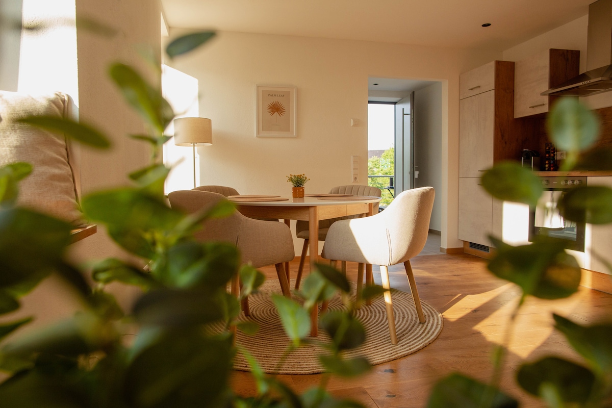 A dining area is visible, featuring a round wooden table surrounded by four curved chairs. Natural light floods the room, highlighting a small centerpiece plant on the table. In the background, a kitchenette can be glimpsed, with a door leading to an outdoor space.