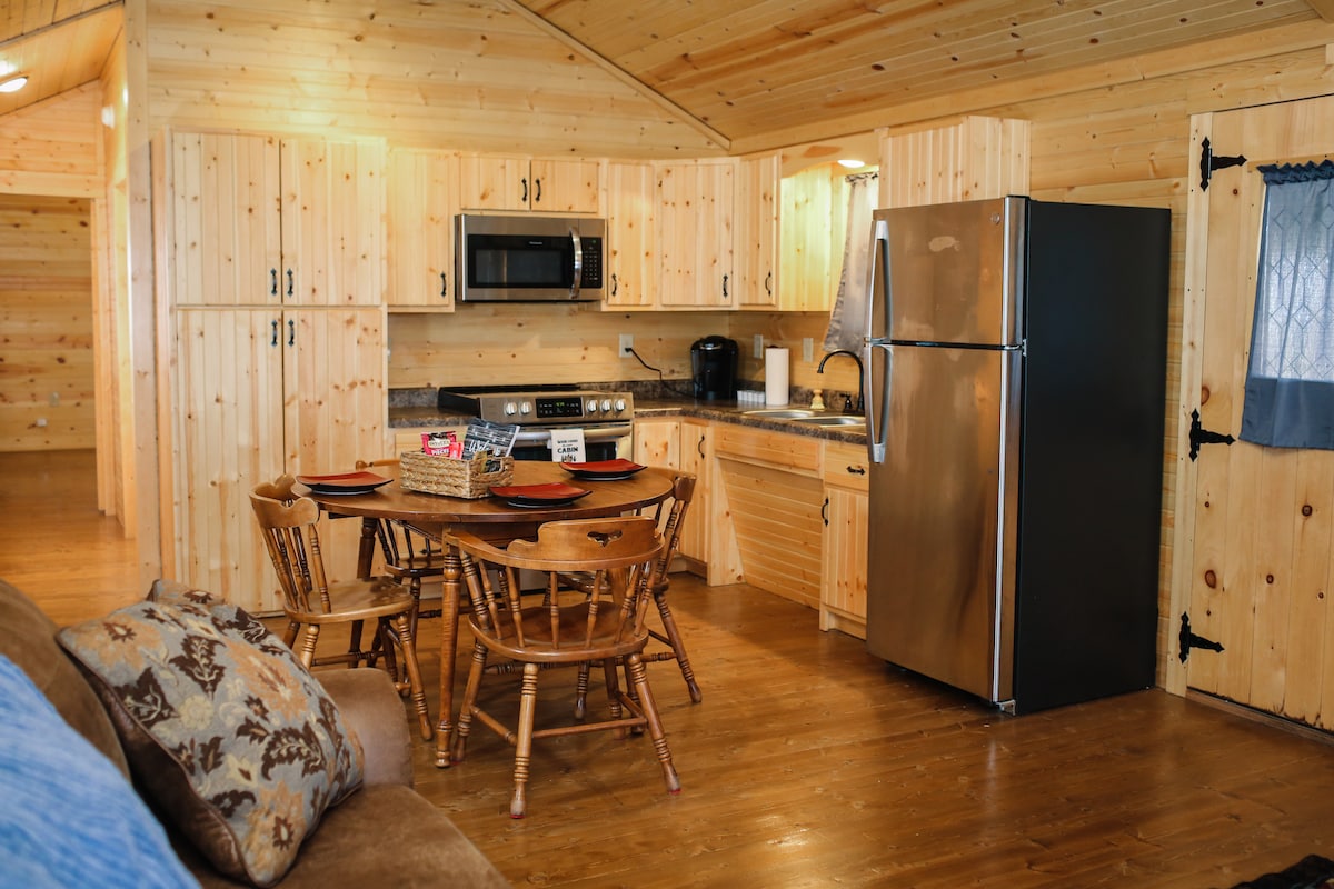 A warm cabin kitchen area is visible, featuring wooden cabinetry and numerous appliances, including a refrigerator, microwave, and stove. A wooden dining table with chairs sits at the center. The space has a rustic charm, highlighted by natural wood paneling on the walls.