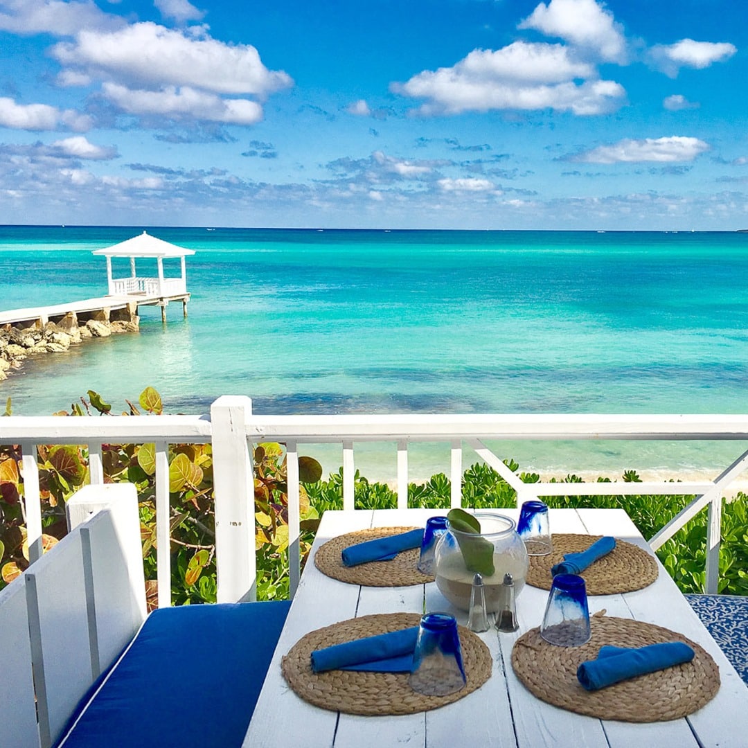 A dining table set for four is positioned on a terrace overlooking a tranquil turquoise ocean. Blue napkins are arranged on woven placemats, while a pitcher and glasses sit at the center. A white gazebo extends over the water in the distance, framed by lush greenery.
