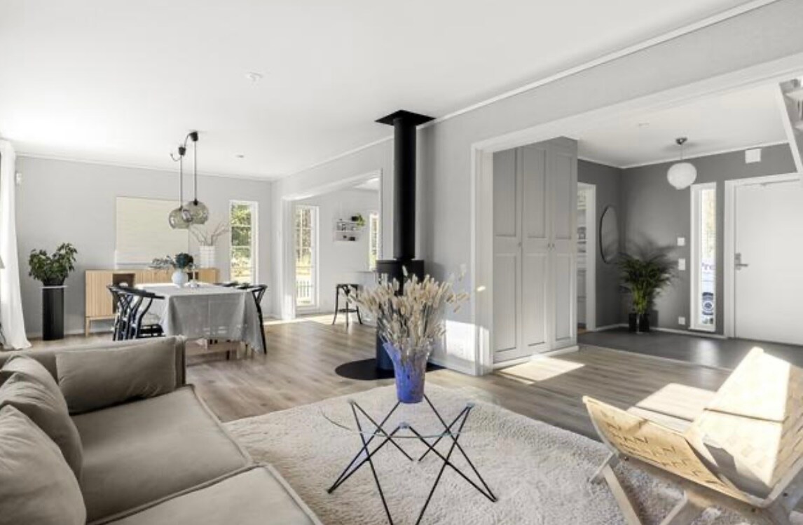 An open living space features a comfortable sofa paired with a modern coffee table. A dining area with elegant furnishings is visible, along with a cozy braskamin. Natural light floods through large windows, enhancing the warmth of the light-toned flooring and walls.