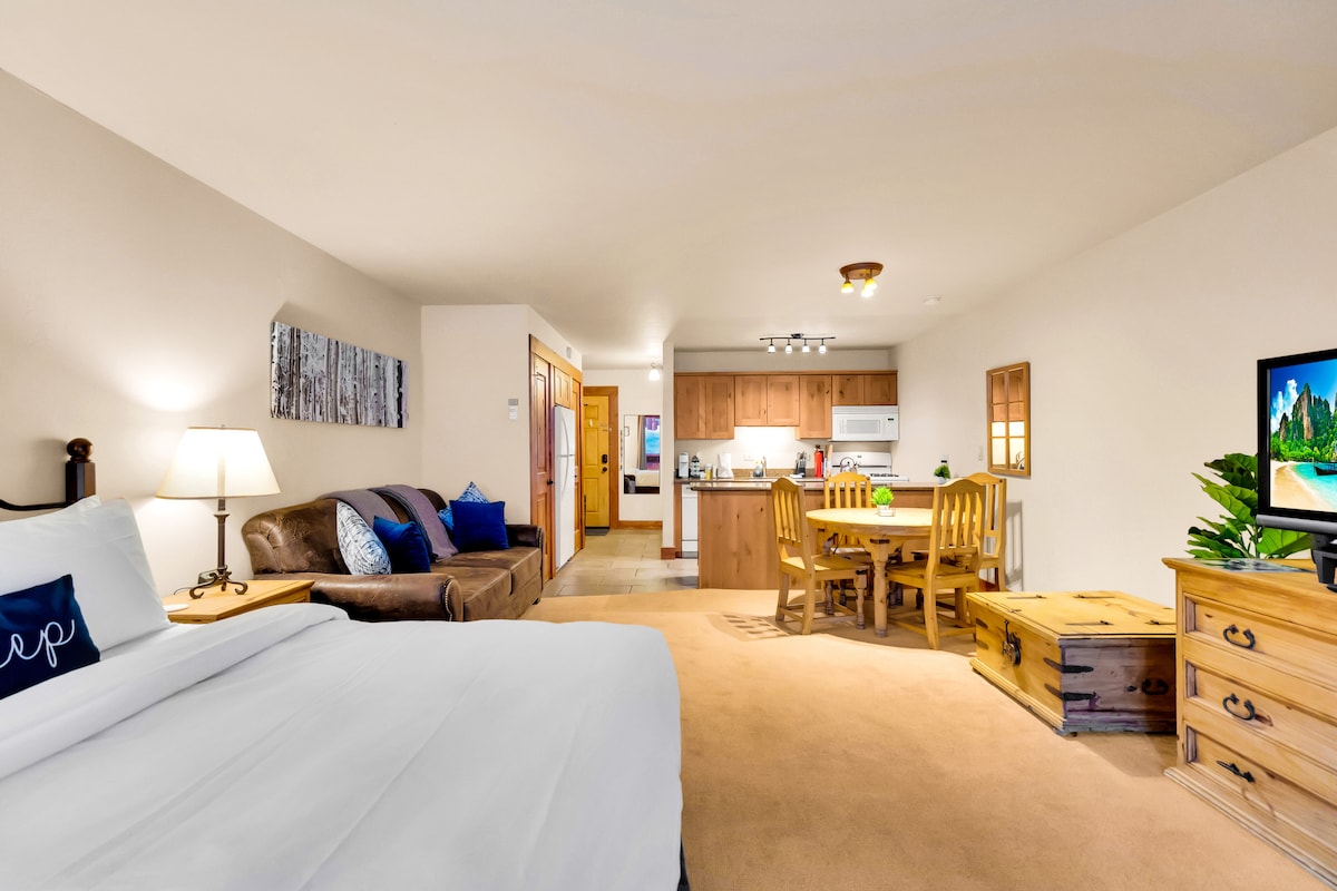 A spacious room features a comfortable bed beside a lamp and a dark couch. A dining area with wooden chairs surrounds a table, while an open kitchen with appliances is visible in the background. Natural light brightens the area, creating a welcoming atmosphere.