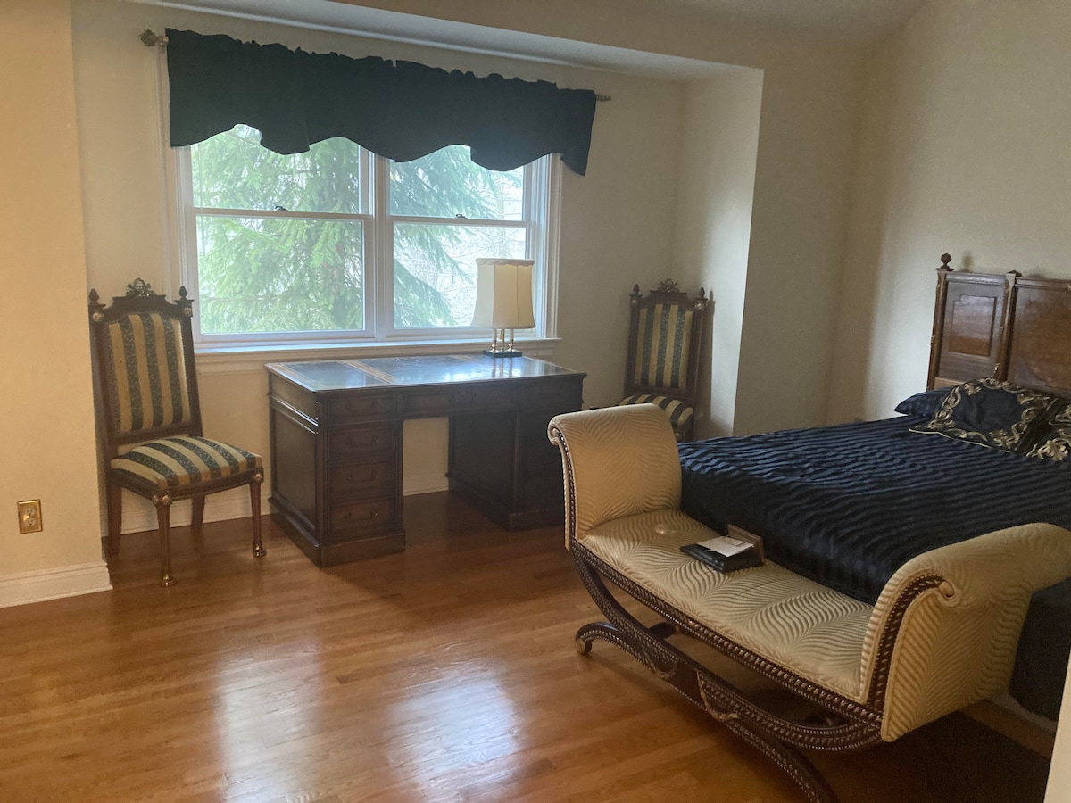 A serene bedroom features a spacious bed with dark bedding. Two elegant chairs flank a wooden desk near a large window draped with a dark valance. Natural light enters the room, illuminating the polished wooden floor.