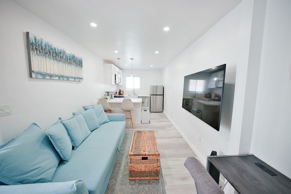 A light-filled living area features a soft blue couch paired with neutral seating. A wooden storage basket is positioned in front. The view extends to a modern kitchen equipped with stainless steel appliances and bright countertops, illuminated by recessed lighting and a window.