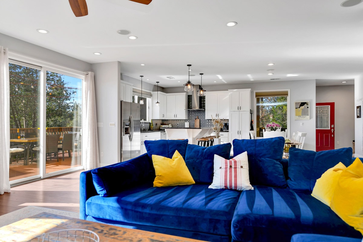 A spacious living area features a large blue sectional couch adorned with bright yellow pillows. A modern kitchen is visible in the background, complemented by white cabinetry and pendant lighting. Floor-to-ceiling sliding glass doors open to a deck, inviting natural light into the space.