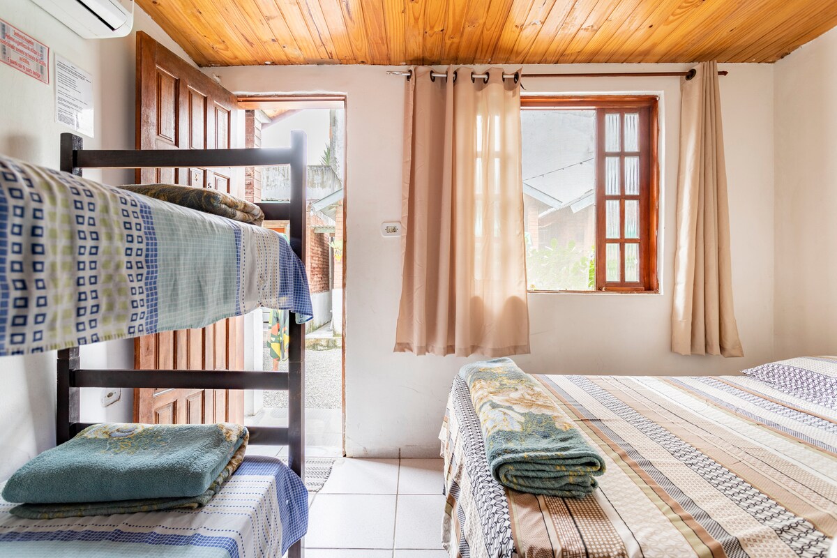 A bright sleeping area features a spacious double bed alongside a wooden bunk bed. Natural light enters through two windows, framed by light curtains. A door leads to an outdoor space, adding to the openness of the room.