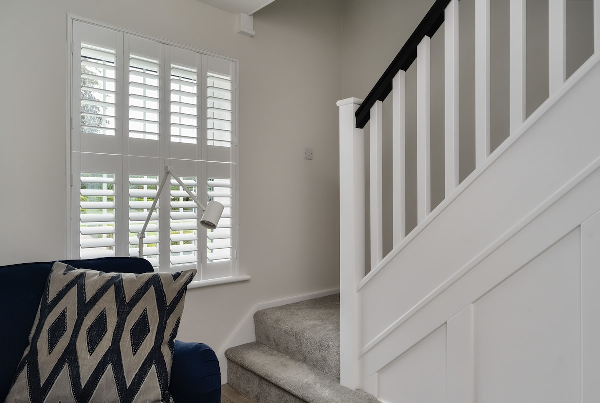 A well-lit corner of the living area showcases a blue sofa adorned with a patterned throw pillow. Window shutters allow natural light to filter in. The staircase, with its white railings, leads to the upper level, enhancing the space's openness and inviting atmosphere.