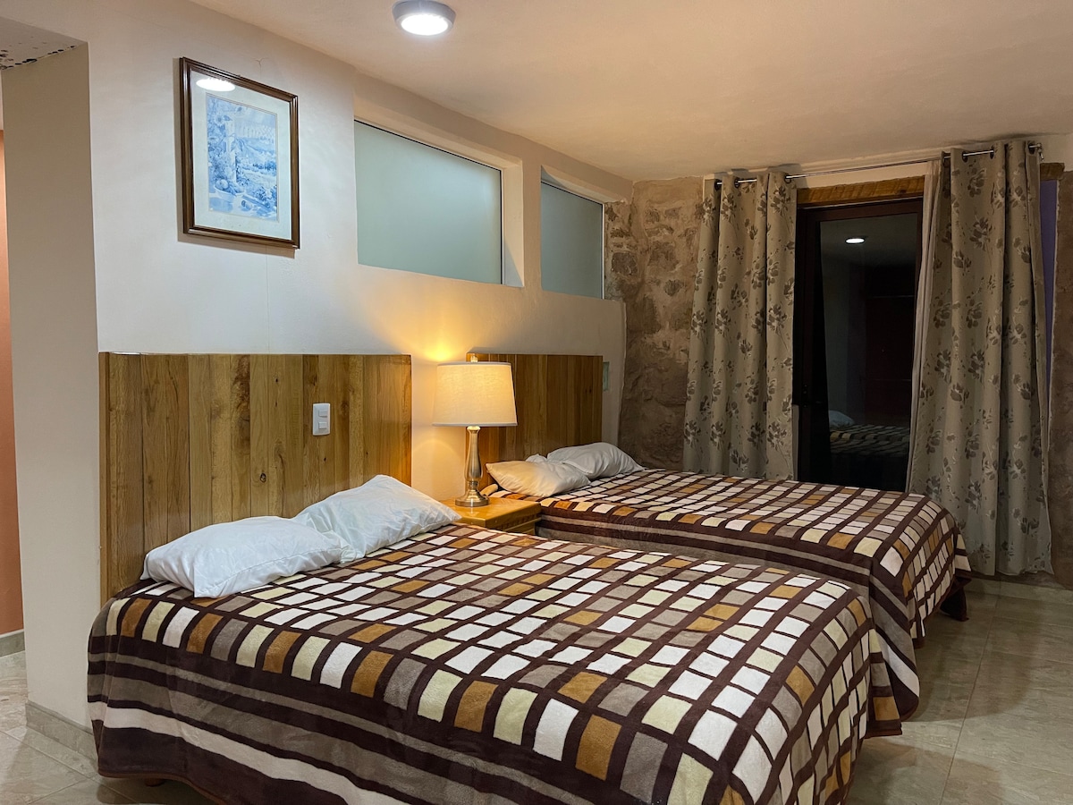 A well-lit sleeping area features two spacious beds with patterned bedspreads. Soft lighting is provided by a bedside lamp, and light-colored curtains frame the windows. A doorway leads to an outdoor space, adding to the bright, welcoming environment.