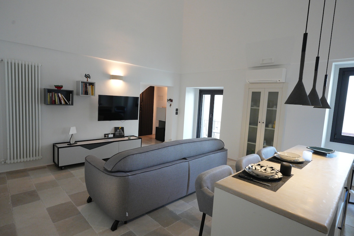 The open living space features a gray sofa positioned against a wall, complemented by a sleek television unit. A dining area with minimalist décor includes a kitchen island adorned with stylish tableware. Large windows invite natural light into the neutral-toned room, contributing to a welcoming environment.