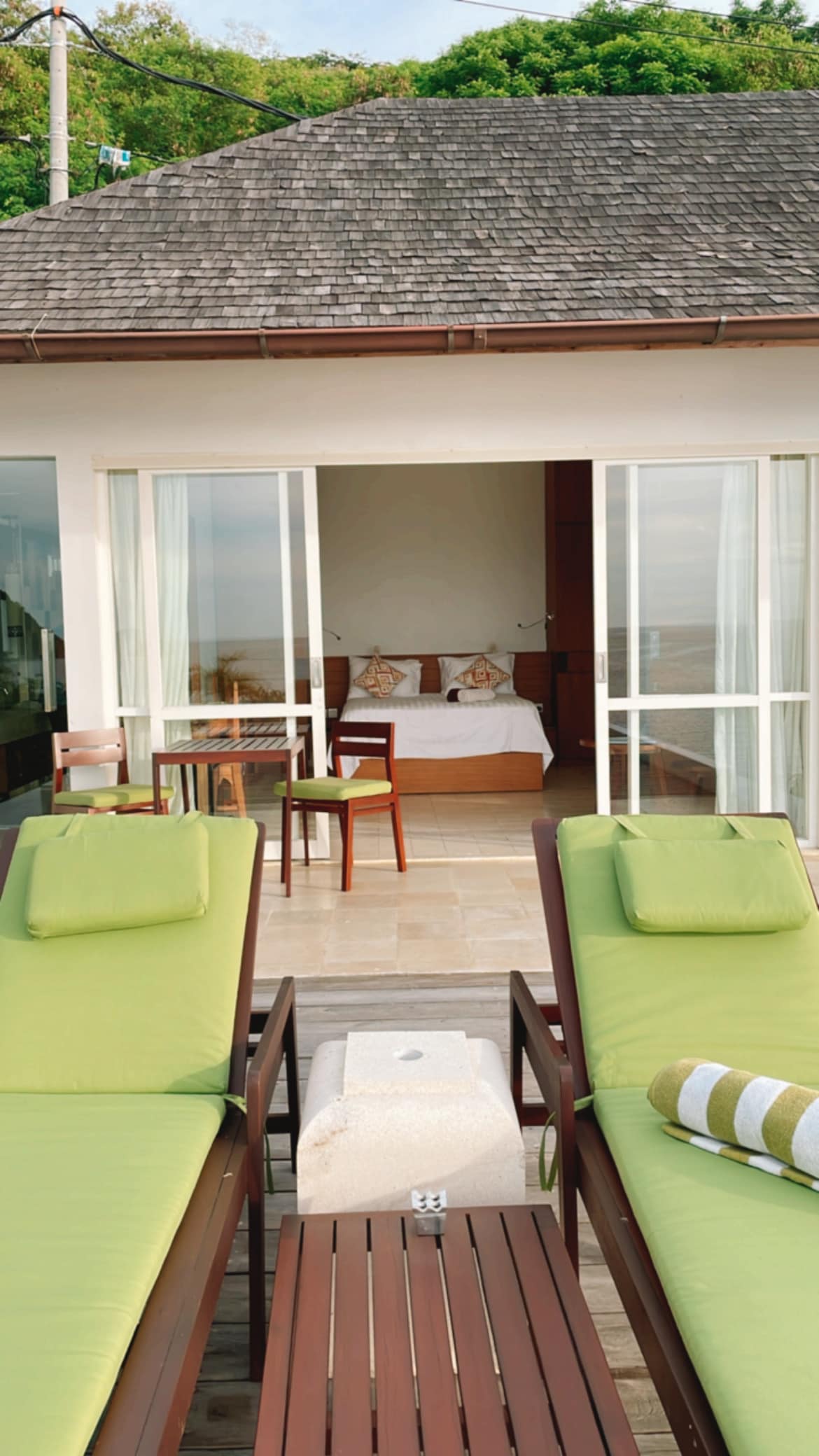 Two comfortable lounge chairs with green cushions are arranged on a stone patio, facing large glass doors that open to a bright bedroom. A small table sits between the chairs, while a rolled towel adds a decorative touch.