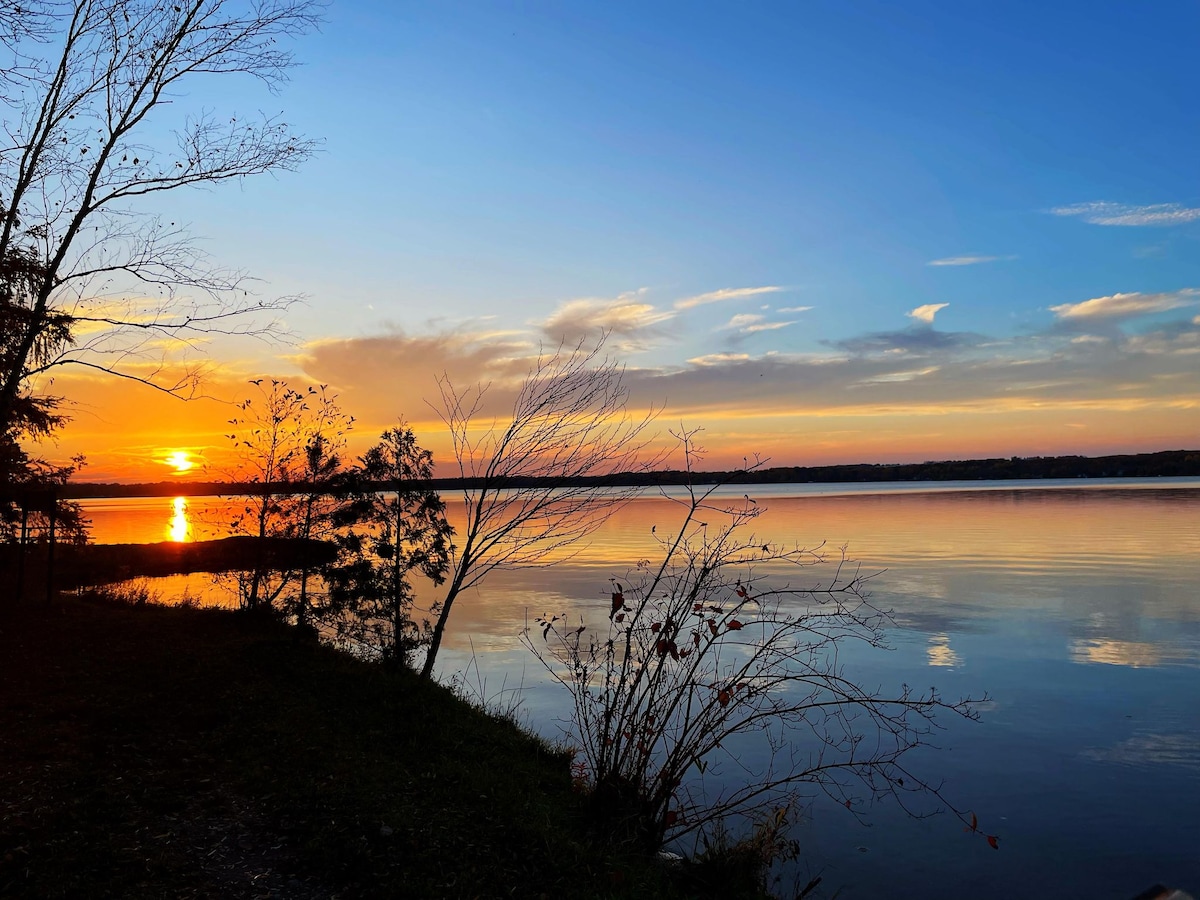 A serene lakeside view captures the sunset reflecting on the calm water. Trees along the shore are silhouetted against the colorful sky, which transitions from golden hues to deep blue. The stillness of the lake enhances the tranquil atmosphere of the setting.