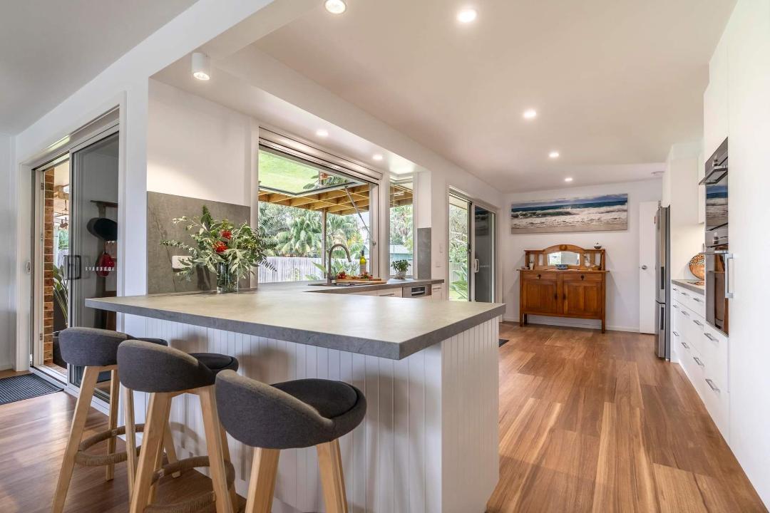 A modern kitchen is presented with a central island featuring four bar stools. Large windows allow natural light to fill the space, illuminating the sleek cabinetry and contemporary appliances. An inviting view of the surrounding greenery can be seen through the open doors.