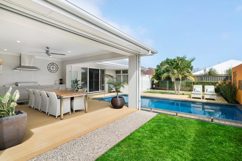 An open-plan indoor and outdoor living space showcases a dining area with a large table and multiple chairs. A solar heated pool is visible alongside a lush green lawn, framed by palm trees. A seating area is also present near the pool, enhancing the relaxing environment.
