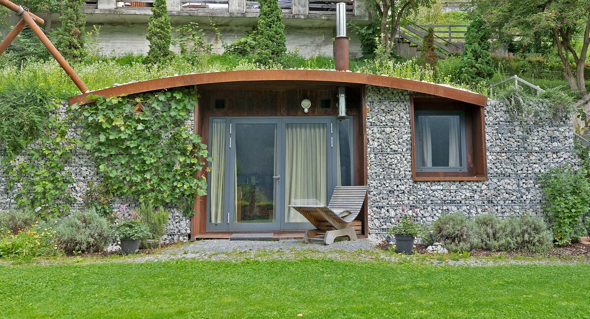 The entrance to the earth house is framed by lush greenery and stone accents. Large glass doors invite natural light into the space. A wood chair sits on a grassy area, providing a spot to relax amidst the surrounding landscape.