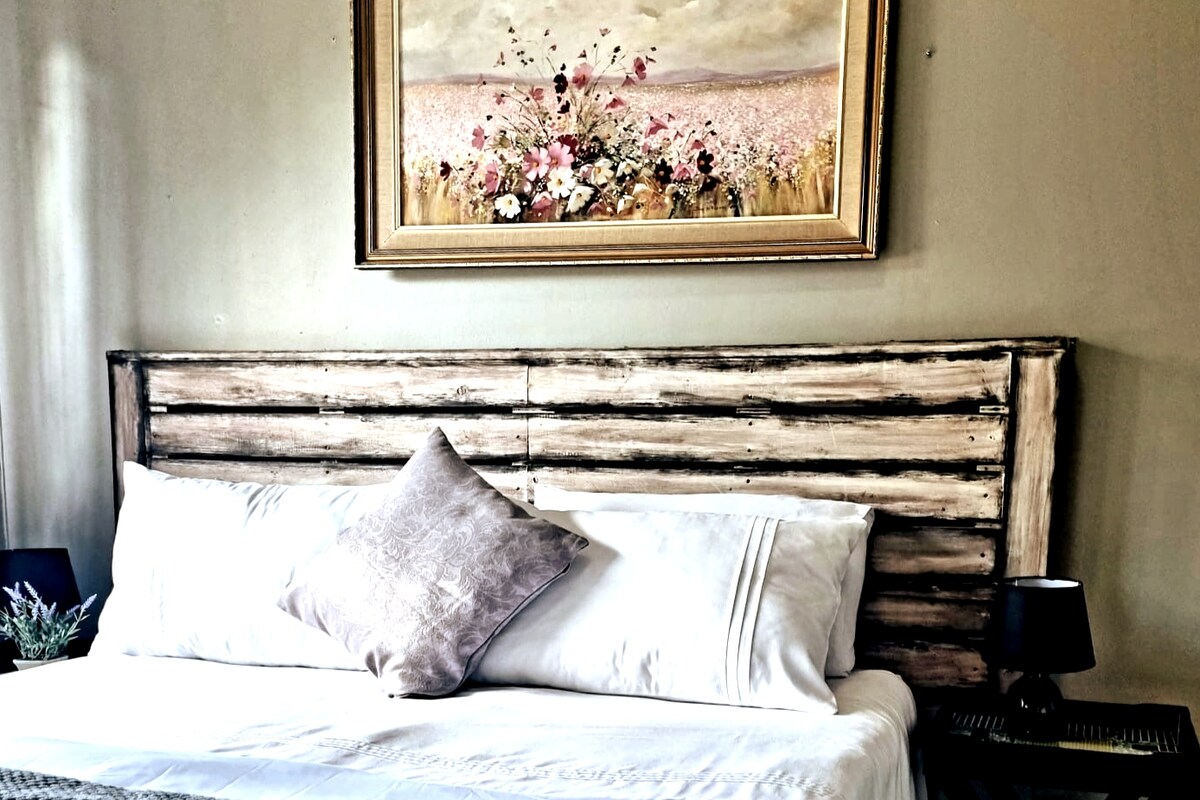A spacious bed is positioned against a wooden headboard with a rustic design. Above the bed, a framed painting featuring a floral arrangement provides a touch of color against the neutral wall. Soft bedding is complemented by a decorative pillow and matching bedside lamps.