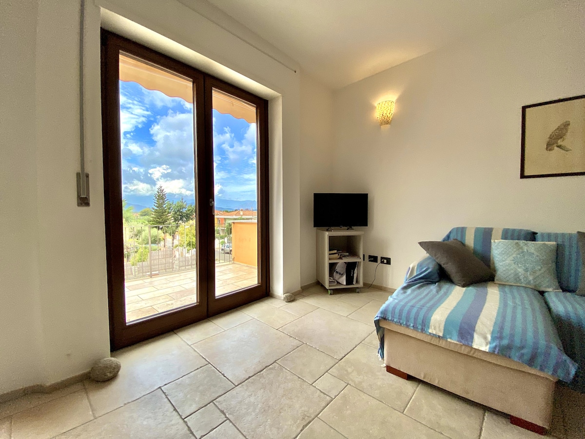 A bright living area is showcased with large glass doors leading to a balcony, revealing views of the surrounding landscape. A comfortable sofa with blue and white stripes is positioned in the space, accompanied by a small television and soft lighting from a wall fixture.
