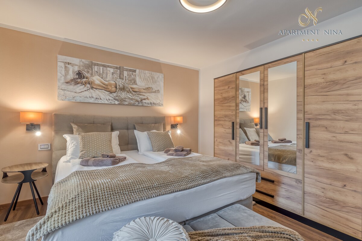 A spacious bedroom features a comfortable bed with neutral bedding, flanked by soft lighting. A large mirror reflects the decor, and a piece of wall art adds a touch of elegance. A bedside table holds essentials, enhancing the warm ambiance of the room.