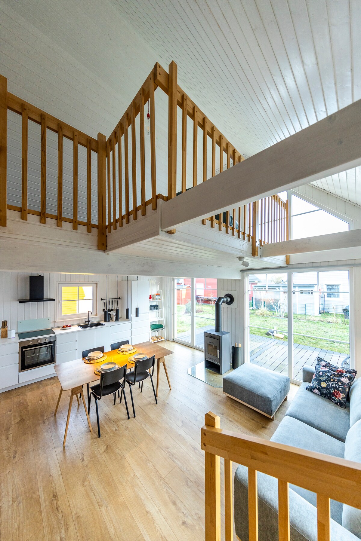 A spacious living area features high ceilings with wooden beams and large windows that offer views of the garden and harbor. The modern kitchen includes sleek cabinetry and a dining table set for four, while a cozy sofa is positioned near a stylish wood stove.