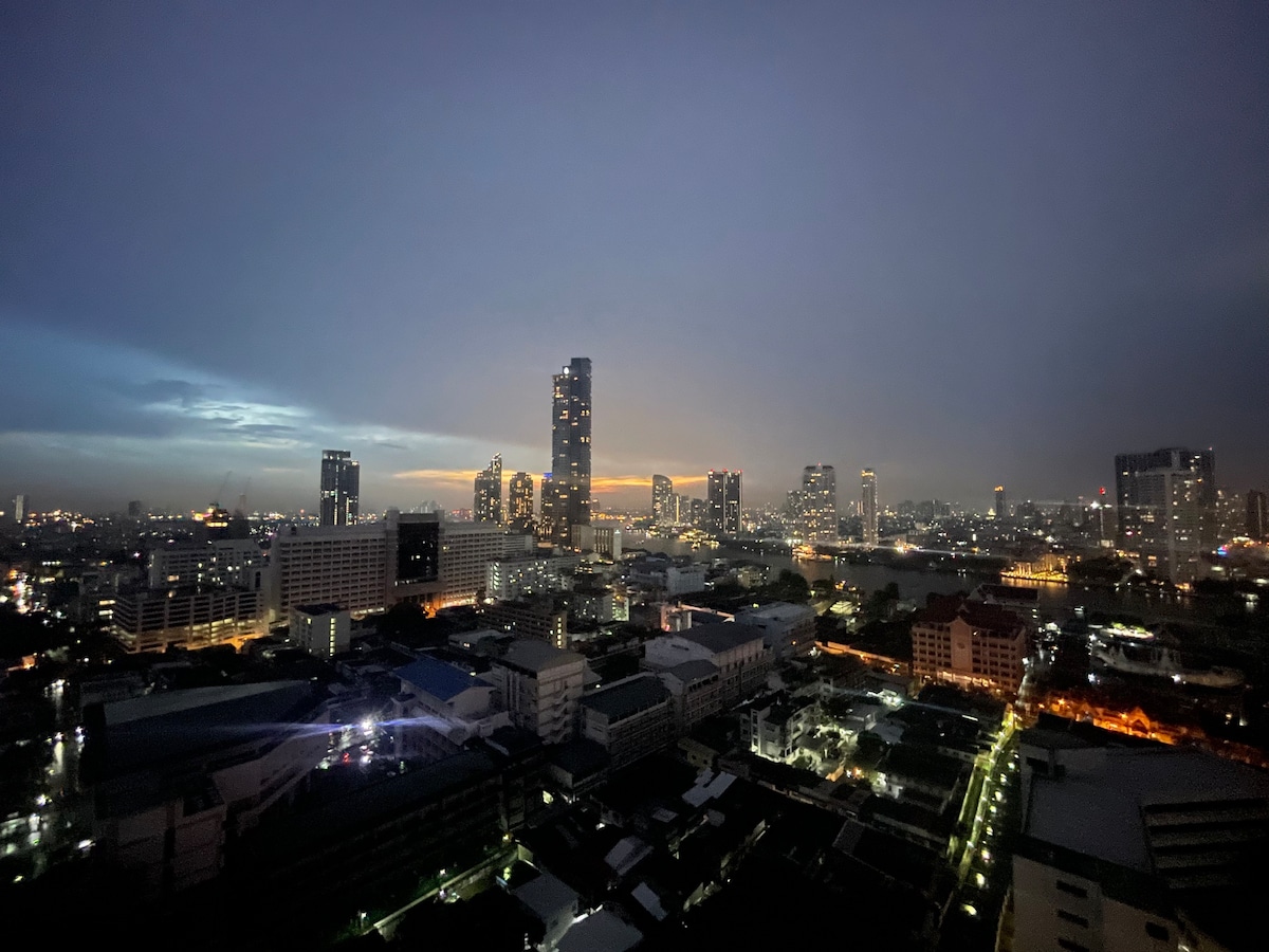 A panoramic view of a city skyline is captured at dusk, showcasing a mix of tall buildings and low-rise structures. The horizon is painted with soft hues of orange and blue, while city lights begin to twinkle as the sky darkens.